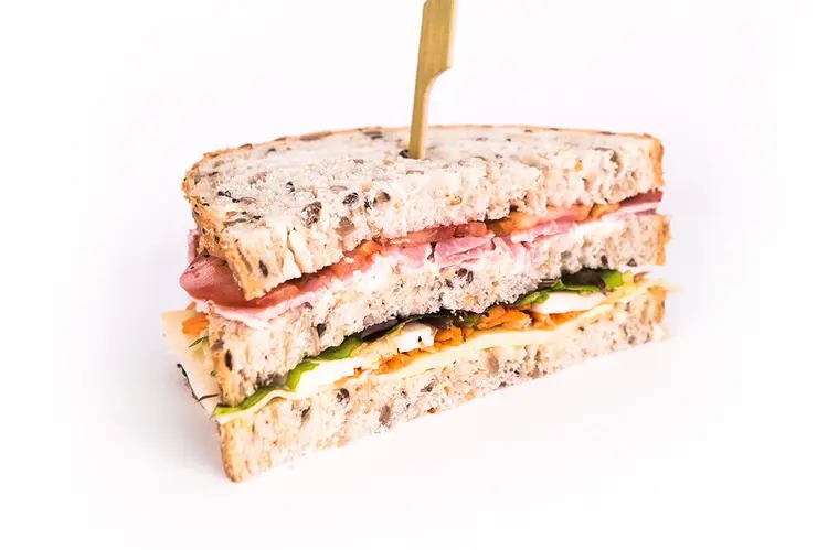 Sandwich Club Bien Belge sur pain aux graines, trois tranches garnies de jambon, fromage et roquette, maintenu par une brochette en bambou