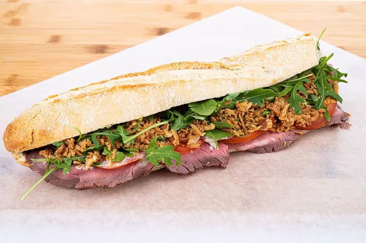 Sandwich La Nonna ouvert sur baguette artisanale — rosbif saignant, oignons frits croustillants, roquette et tranches de tomate