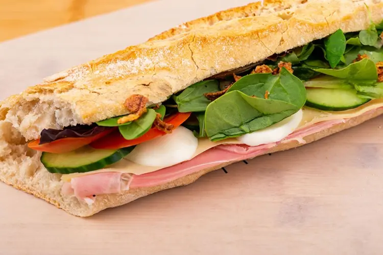 Baguette gourmande Tartineke ouverte sur planche en bois — jambon, mozzarella, tomate, concombre, épinards et oignons frits — fabriquée au centre de Wavre